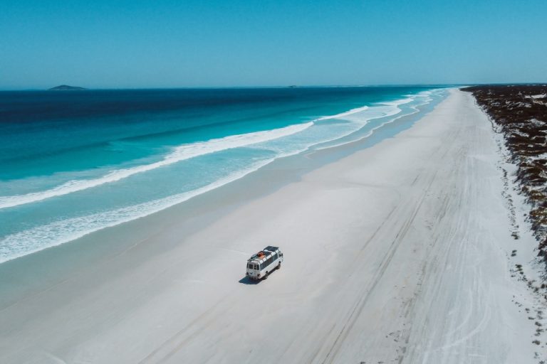 A white van driving along the vast, pristine shoreline of Cape Le Grand Beach with turquoise waves and a clear blue sky, one of the many things to do in Western Australia
