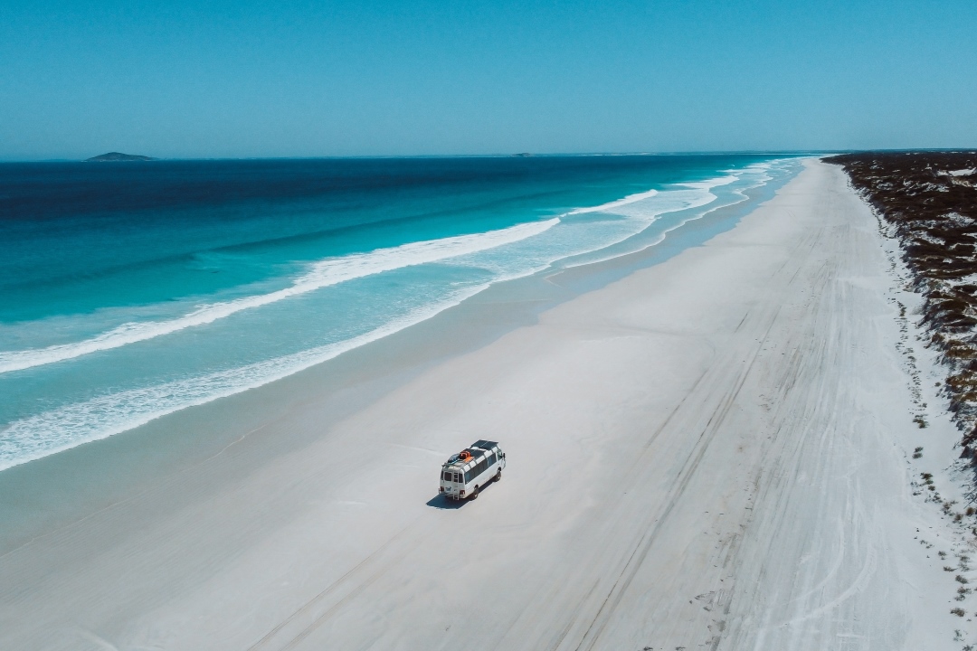 A white van driving along the vast, pristine shoreline of Cape Le Grand Beach with turquoise waves and a clear blue sky, one of the many things to do in Western Australia
