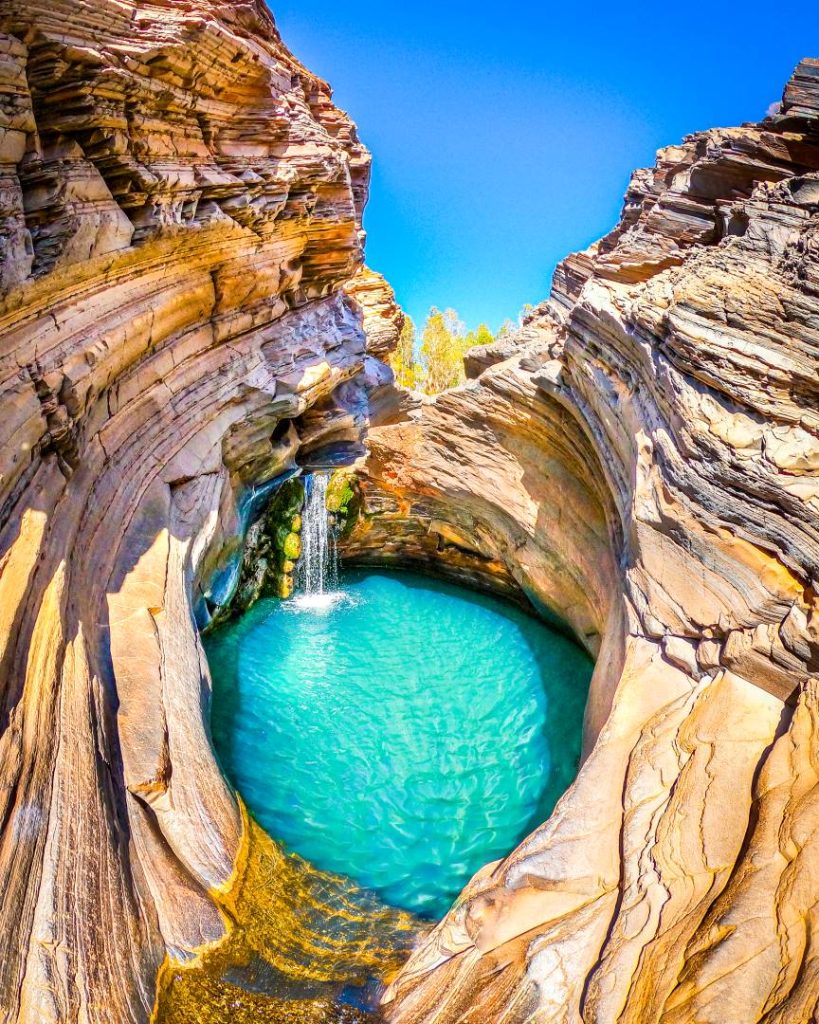 The turquoise waters of Spa Pool, a natural rock pool with a small waterfall and curved rock walls in Hamersley Gorge, one of the best gorges in Karijini. The sunlight highlights the layered rock textures.