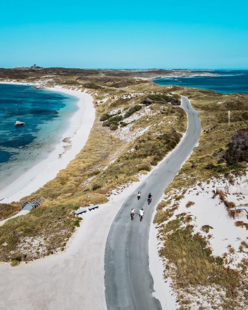 Cyclists riding along a winding coastal road with turquoise waters and sandy dunes on Rottnest Island, one of the many things to do in Western Australia
