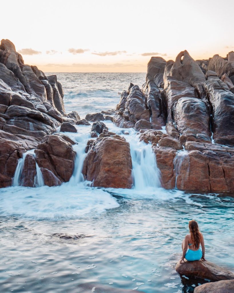 A woman sitting by a rock pool with waves crashing through the Injidup Natural Spa at sunrise, a popular thing to do in Western Australia.