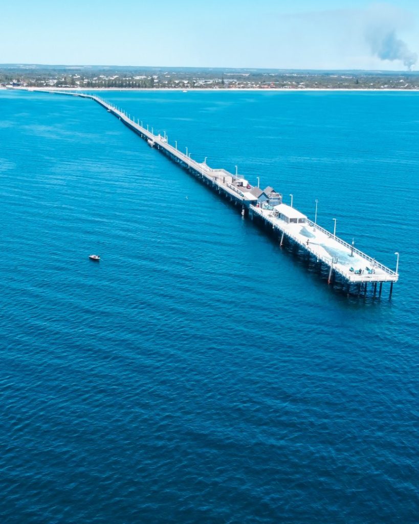 A long white jetty extending into turquoise waters with the shoreline in the distance at Busselton.