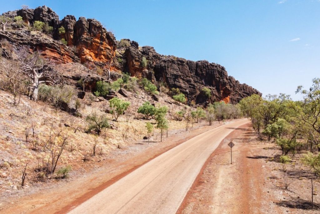 A scenic view of the rugged Gibb River Road, surrounded by rocky cliffs with shades of orange and green vegetation under a clear blue sky.