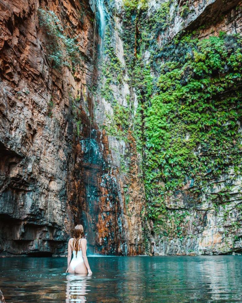 A woman wading in the pristine water of Emma Gorge, surrounded by lush greenery and a dramatic rock wall with a cascading waterfall.