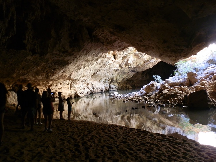 People exploring the illuminated cavern of Tunnel Creek with its reflective water and intricate rock formations.