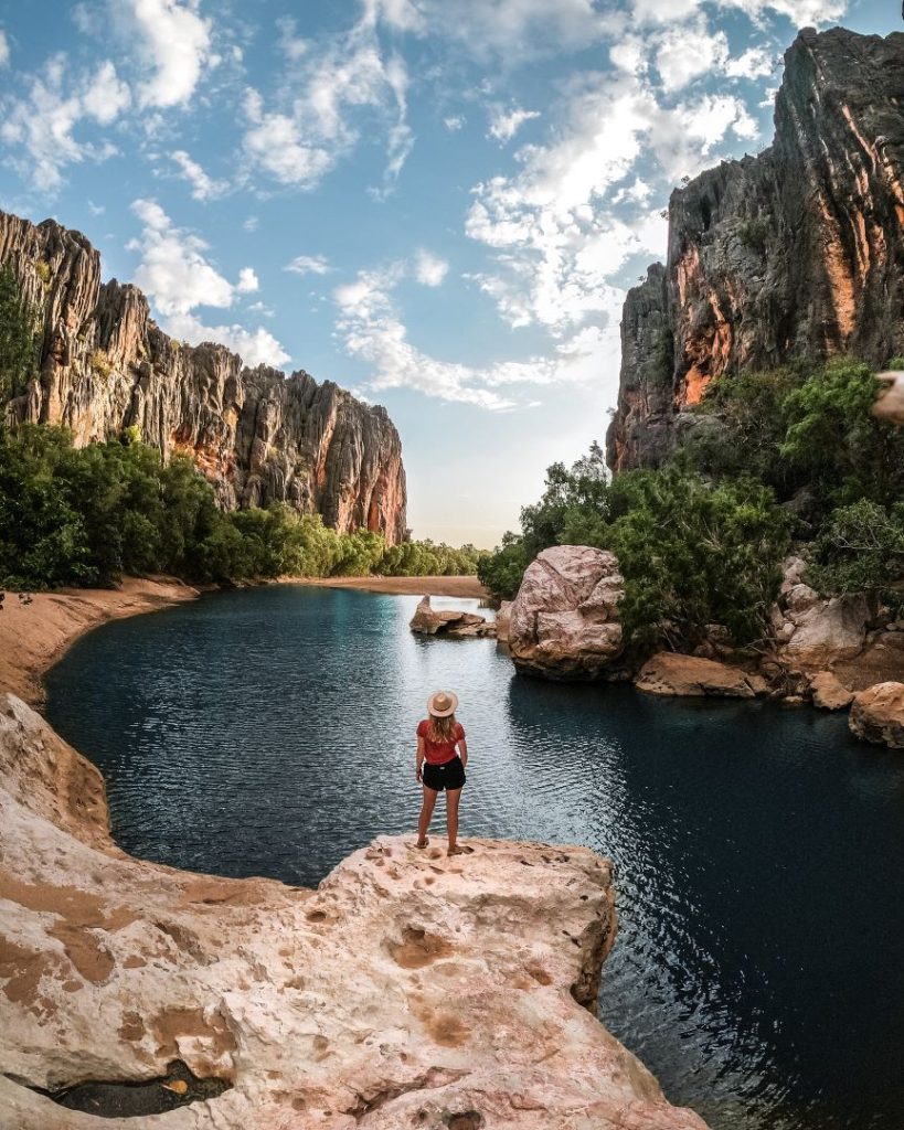 A person standing on a rocky ledge overlooking the calm waters of Windjana Gorge, surrounded by towering cliffs.
