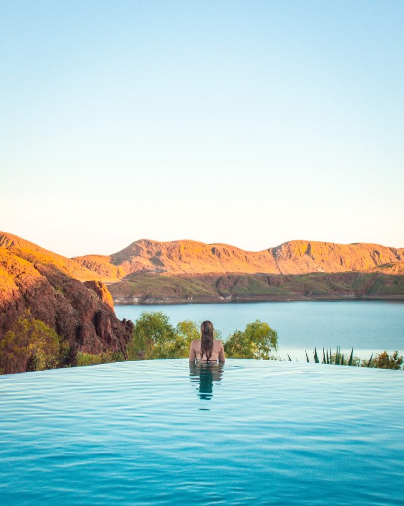 A person enjoying the tranquil infinity pool at Kununurra, overlooking lush landscapes and rugged red cliffs at sunset.