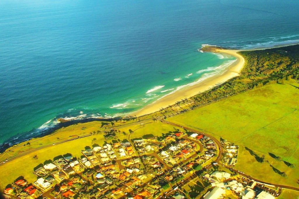 An aerial view of the North New South Wales coastline, showcasing a stretch of sandy beach meeting turquoise waters and a residential area nestled by the coast—a stunning perspective on the scenic things to do in Ballina NSW for those exploring coastal landscapes.