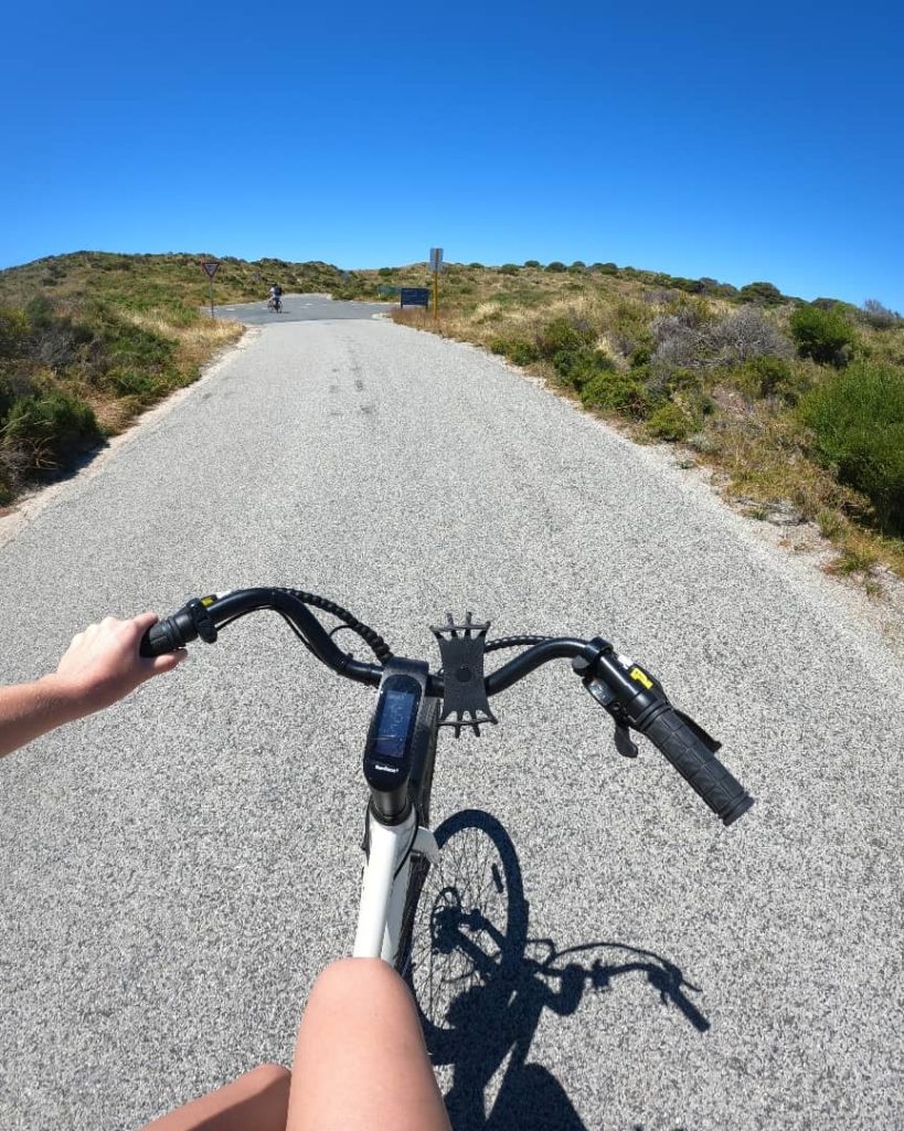 A cyclist's perspective riding a bike on a sunny day along a scenic path on Rottnest Island, surrounded by natural greenery and clear blue skies.