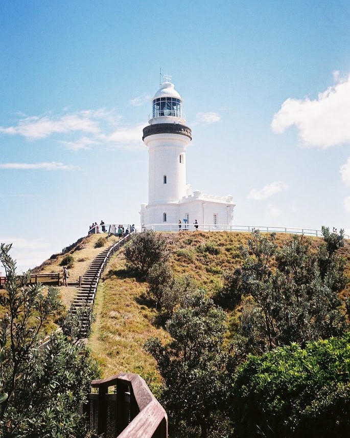 The iconic Cape Byron Lighthouse sits atop a grassy hill under a clear blue sky, with visitors climbing the path to reach its scenic viewpoint—one of the must-visit things to do in Ballina NSW for breathtaking coastal views.