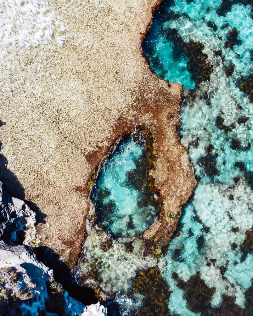 An aerial view of Cape Vlamingh on Rottnest Island, featuring clear turquoise rock pools and rugged coastal terrain surrounded by vibrant reef formations.