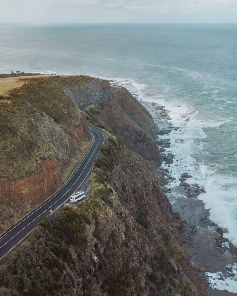 An aerial view of a winding road along the rugged cliffs of the Great Ocean Road in Victoria, Australia. A white van is parked on the roadside, with waves crashing against the rocks below, creating a stunning coastal scene.