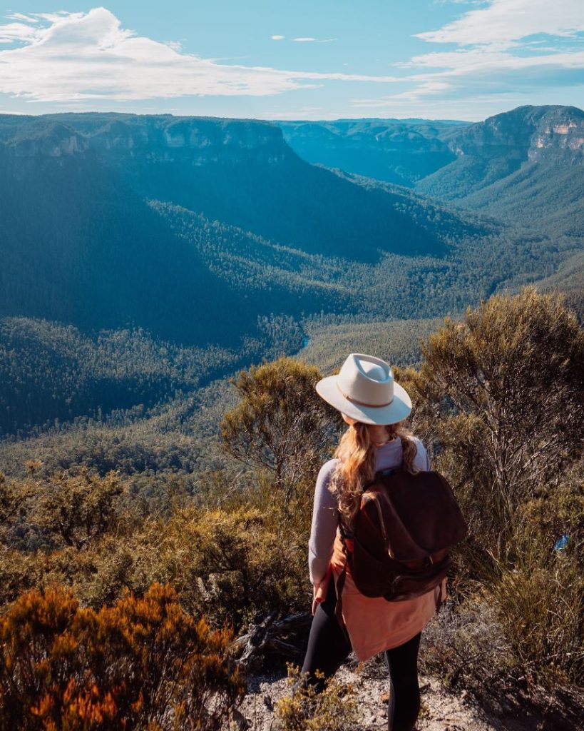 A woman wearing a hat and backpack stands at a scenic lookout in the Blue Mountains, New South Wales, gazing over vast forested valleys and steep cliffs under a bright sky. The rugged landscape highlights the area's natural beauty and appeal for hikers.