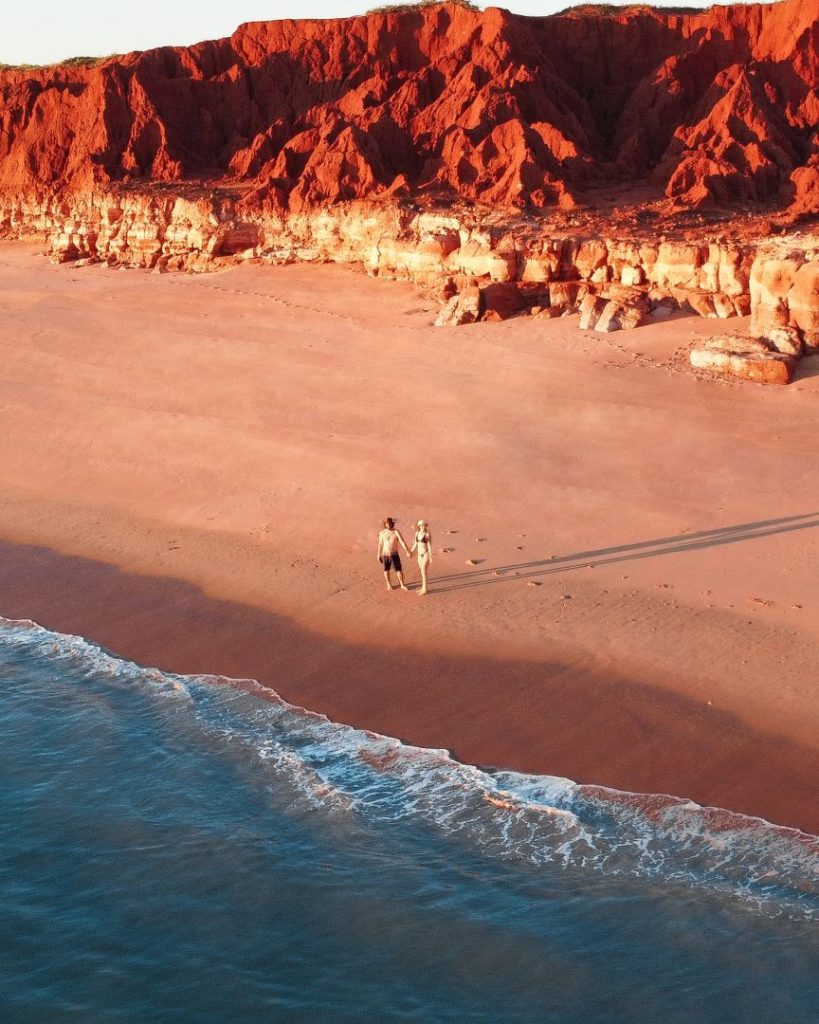 A couple walking hand-in-hand along the shoreline at James Price Point, near Broome, Western Australia. The beach features vivid red cliffs contrasting with the turquoise ocean waves under warm, golden sunlight.