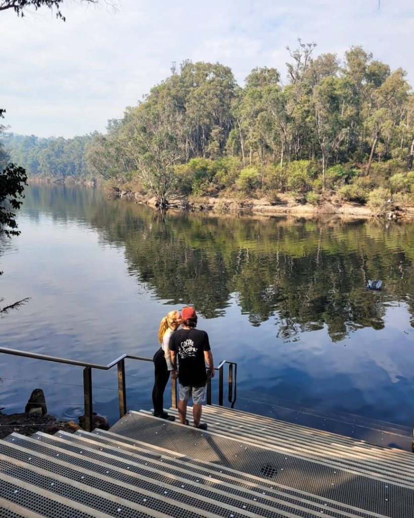 A couple stands on steps leading down to a calm, reflective river surrounded by dense forest at Lane Poole Reserve, a peaceful outdoor destination in Dwellingup.