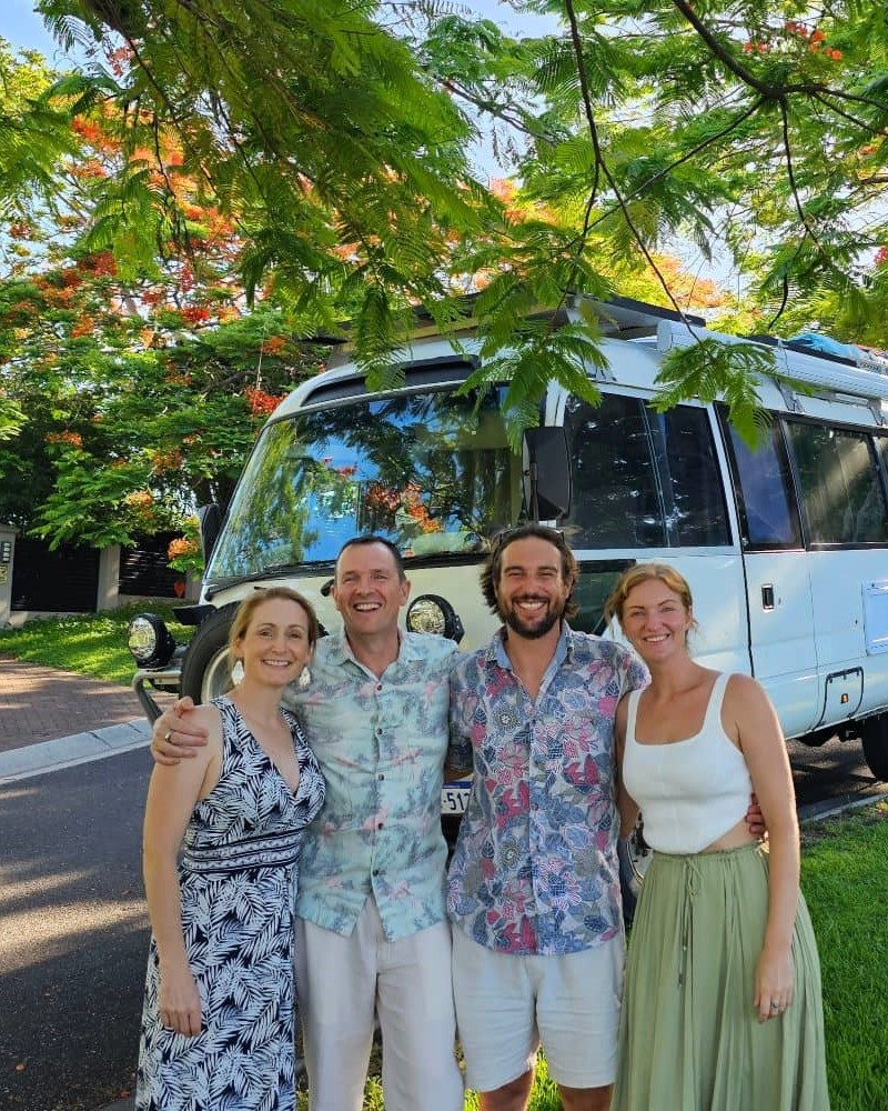 Four smiling adults pose arm-in-arm in front of a white motorhome under a tree with bright green leaves and red-orange blossoms