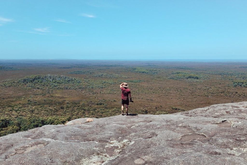 Hiker on the summit of Mount Chudalup in Windy Harbour WA, overlooking a vast wilderness of green forest stretching to the horizon under a clear blue sky.