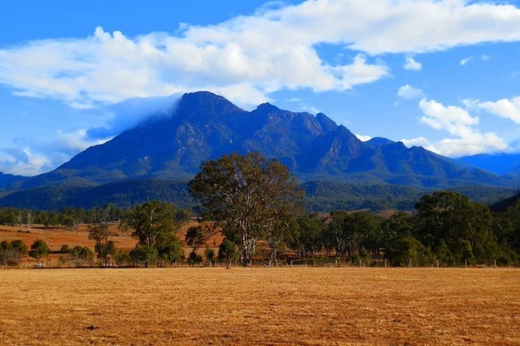 View of Mount Barney's rugged profile against a partly cloudy sky, surrounded by dry fields and scattered trees in the Scenic Rim.