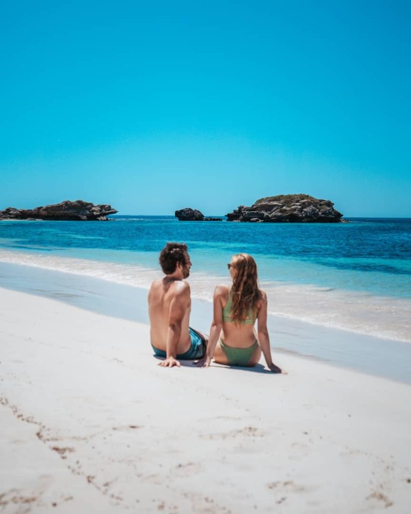 A couple relaxes on the pristine white sands of a beach on Rottnest Island, gazing at the turquoise waters and the rocky Parakeet Island in the distance under a bright blue sky.