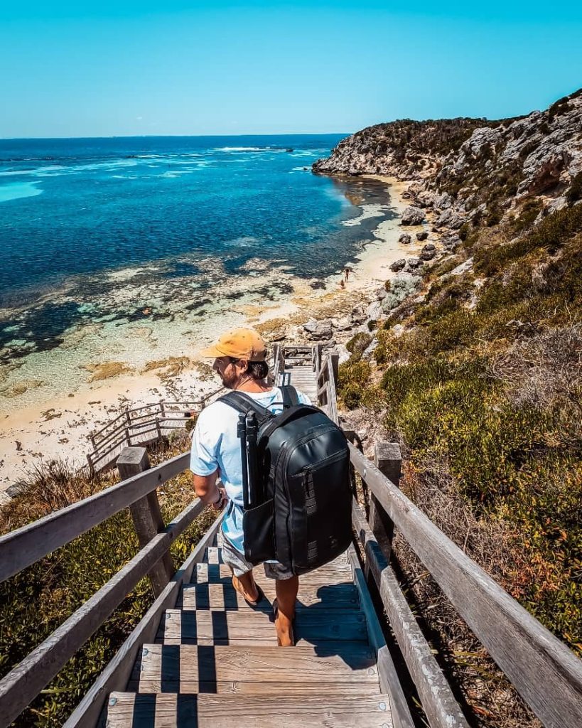 A traveler with a backpack walks down wooden stairs toward the turquoise waters and rocky coastline at Parker Point on Rottnest Island, surrounded by lush vegetation under a bright blue sky.