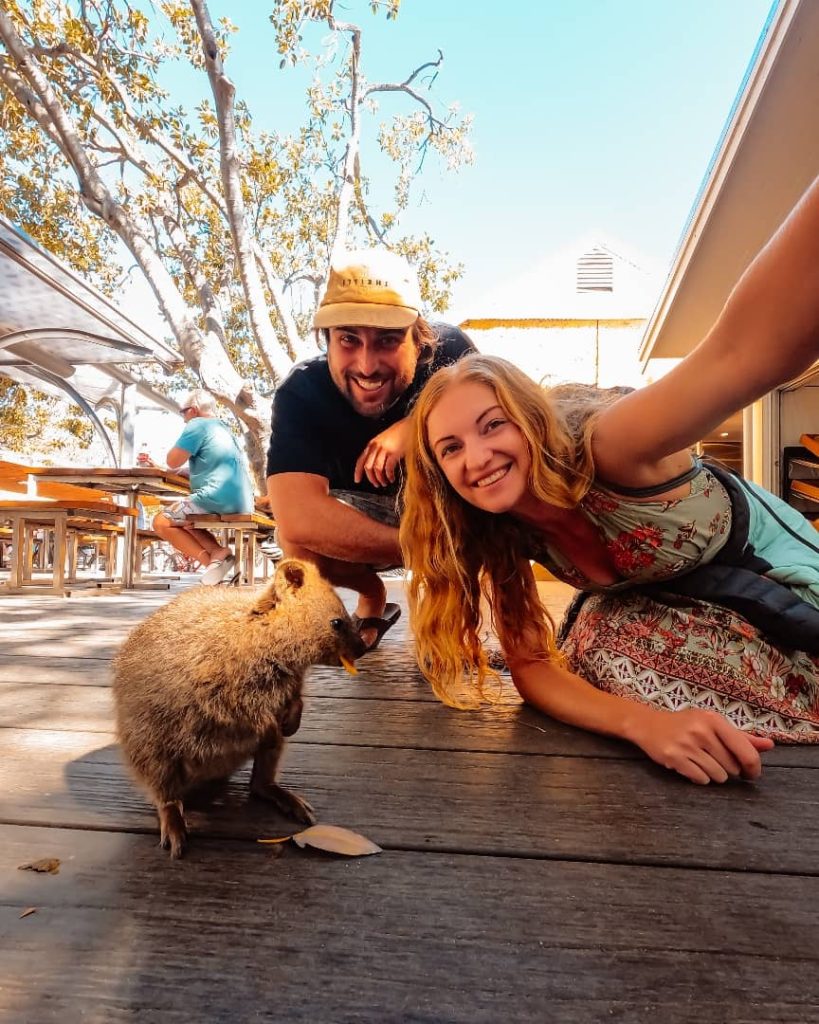 A joyful couple taking a close-up selfie with a friendly quokka on Rottnest Island, showcasing the quokka’s adorable face and playful expression under the shade of trees.