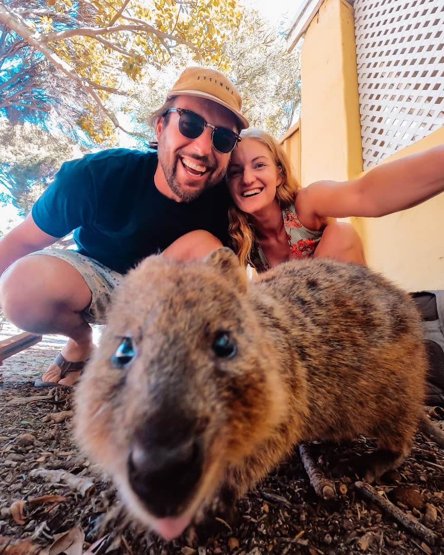 A joyful couple taking a close-up selfie with a friendly quokka on Rottnest Island, showcasing the quokka’s adorable face and playful expression under the shade of trees.