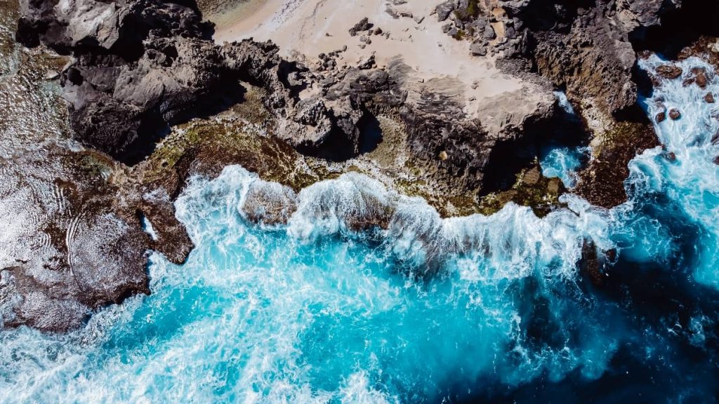 A breathtaking aerial view of waves crashing against rugged rocky cliffs on Rottnest Island, showcasing turquoise waters and dramatic coastal scenery.