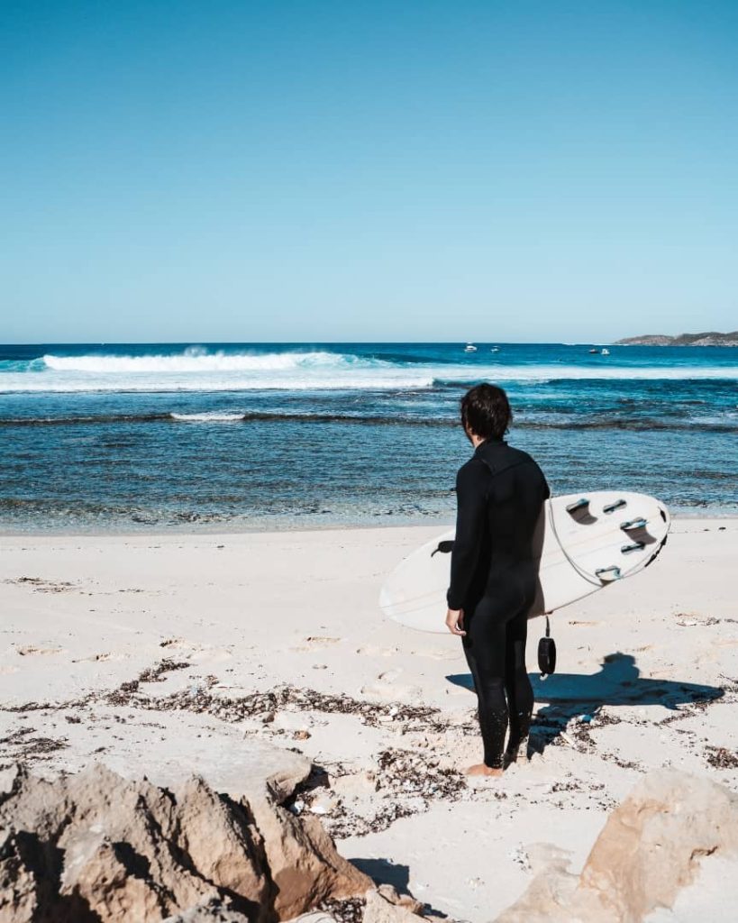A surfer in a wetsuit stands on the sandy beach at Wadjemup, Rottnest Island, holding a surfboard and gazing out at the ocean waves under a clear blue sky.