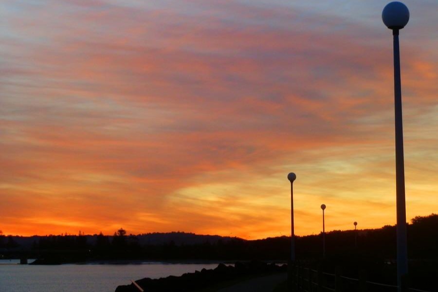 A vibrant sunset casts orange and pink hues over the Richmond River, silhouetting lamp posts along a riverside path in Ballina, New South Wales.