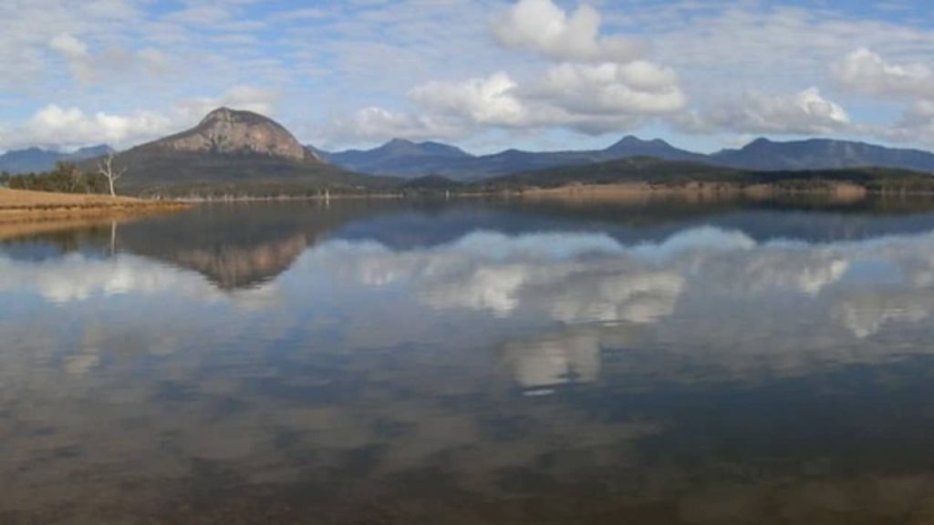 View of the Scenic Rim mountains reflecting in the still waters of Lake Moogerah, Queensland, under a partly cloudy sky.