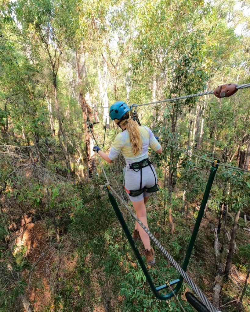A woman wearing a helmet and harness carefully navigates a high rope course among tall trees, showcasing an adventurous activity in Dwellingup’s lush forests.