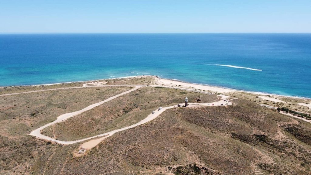 An aerial view of Wadjemup Lighthouse on Rottnest Island, surrounded by rolling hills and pathways, with turquoise ocean waters stretching into the horizon.