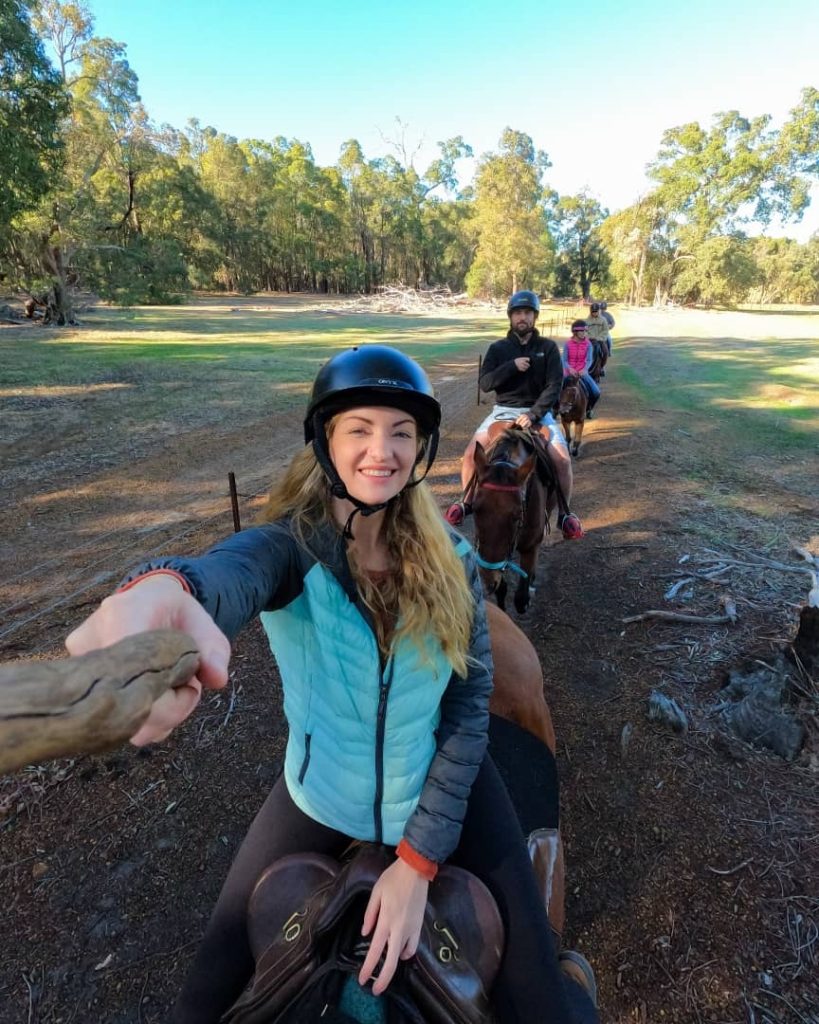 A woman takes a selfie while horseback riding with a group through a scenic, wooded area, showcasing horseback riding.