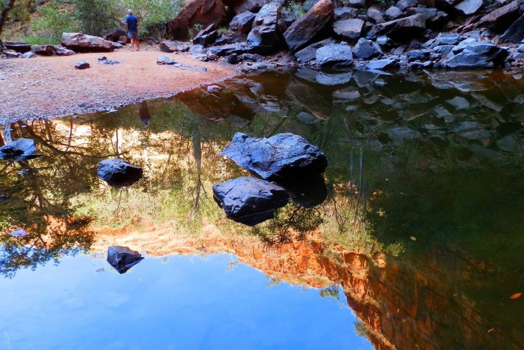 A calm pool at Black Rock in Kununurra reflects the rugged cliffs, scattered rocks, and surrounding greenery, creating a mirrored view of the natural landscape. A lone figure stands on the sandy bank, enhancing the scene’s sense of tranquility and isolation.