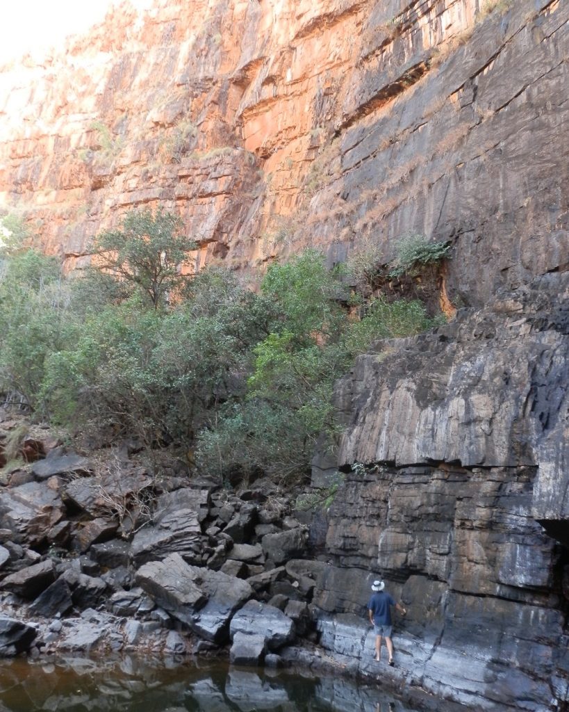 A hiker stands at the base of the towering cliffs at Black Rock in Kununurra, surrounded by rugged rocks and sparse greenery.