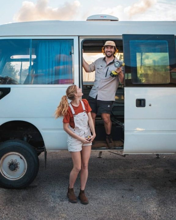 A cheerful couple working on a camper van conversion, with the man standing in the van doorway holding a power tool and the woman looking up at him, smiling. The scene captures a moment of teamwork and excitement during their DIY van build journey.
