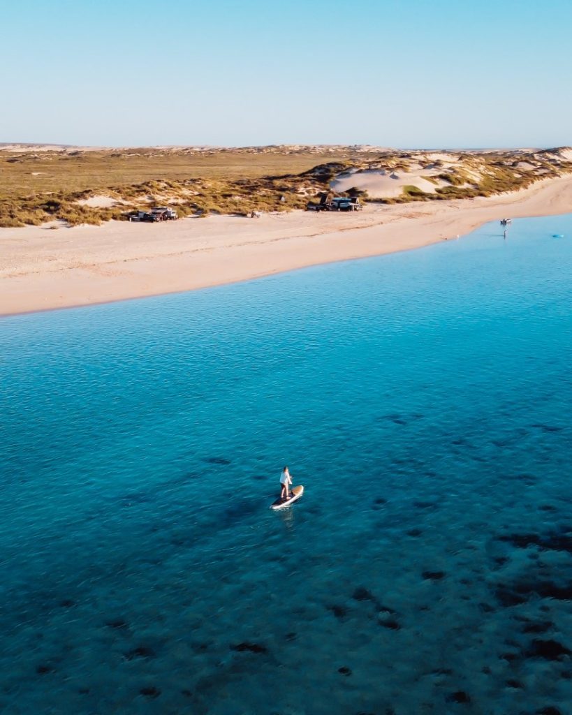 A lone paddleboarder glides over the clear blue waters near a secluded beach, with remote camping vehicles and dunes visible along the shore in the background. This peaceful scene captures the remote beauty of Ningaloo Reef, Australia.