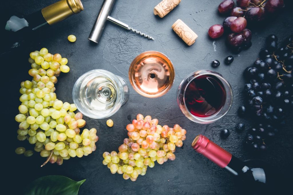 Overhead view of a wine tasting setup featuring three glasses filled with white, rosé, and red wine, surrounded by clusters of green, red, and dark purple grapes. Wine bottles, corks, and a corkscrew are scattered on a dark slate surface, adding to the rustic and inviting arrangement.