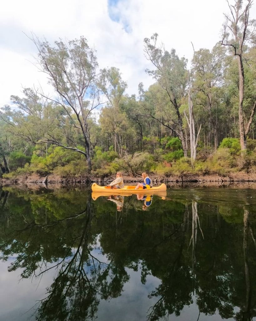 Two people paddle a yellow kayak on a calm river surrounded by dense trees, their reflection mirrored in the still water—a tranquil kayaking experience among the scenic things to do in Dwellingup.