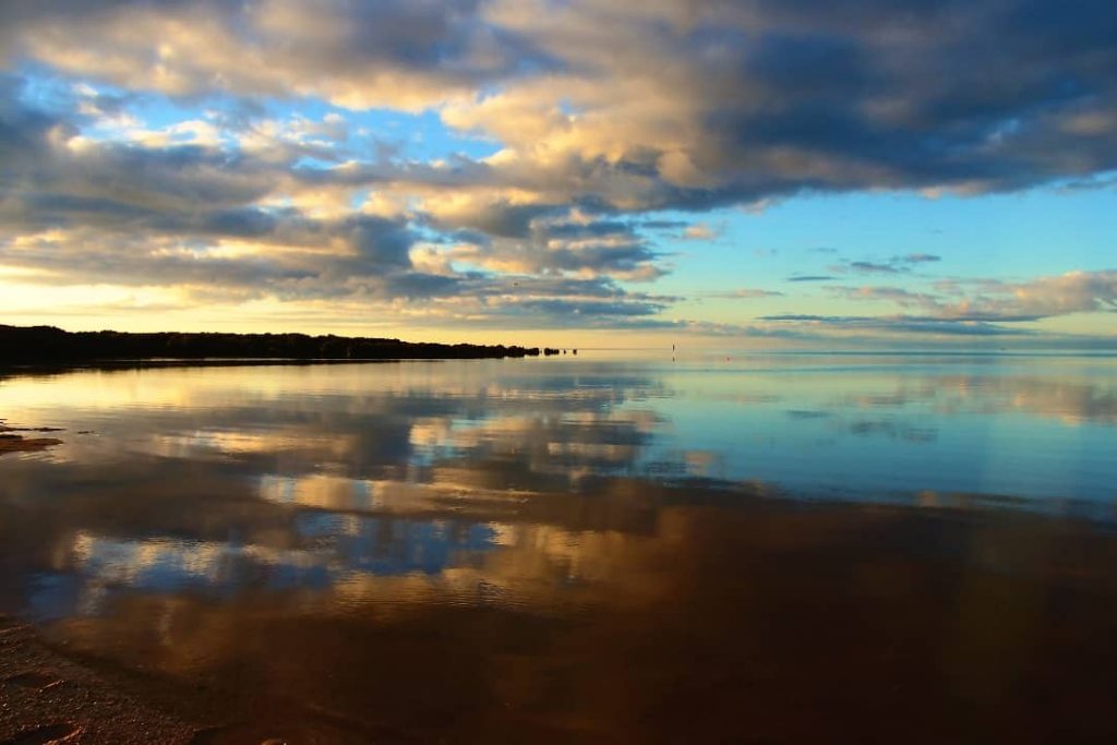 A peaceful morning at Port Clinton, South Australia, with golden sunlight illuminating the sky and clouds beautifully reflected on the calm water's surface.