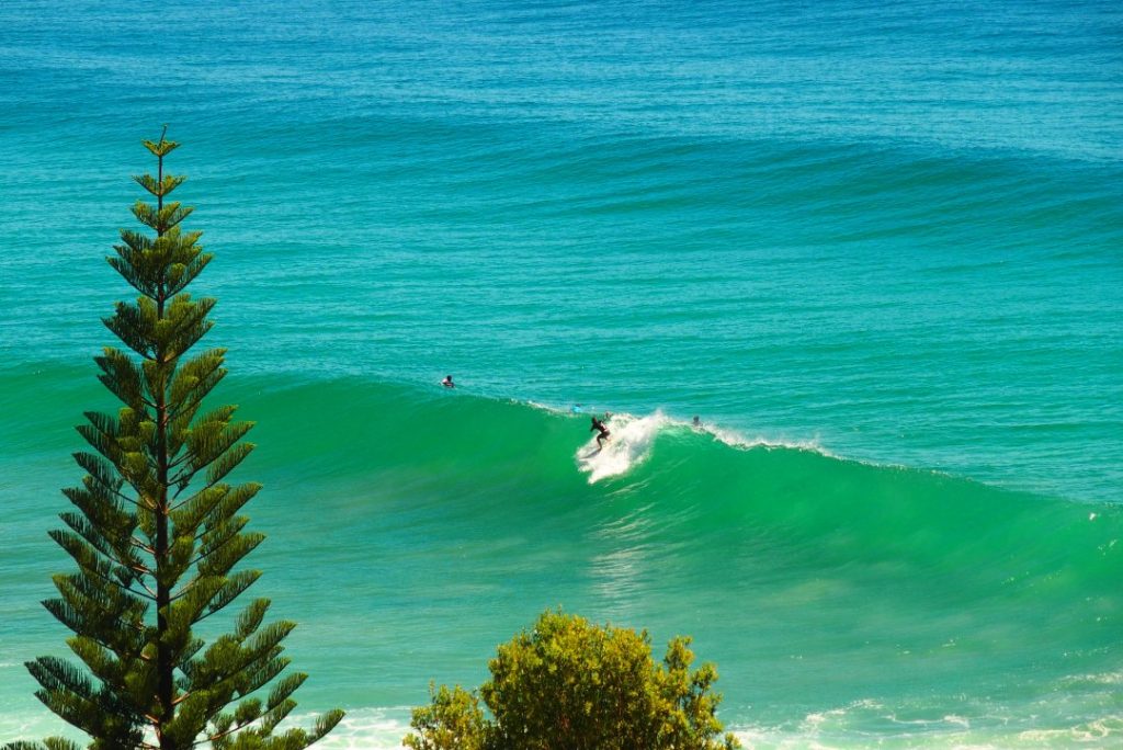 A surfer catches a large wave near the right-hand surf break at Lennox Head, framed by turquoise water and coastal greenery, a popular spot for surfing in Ballina.