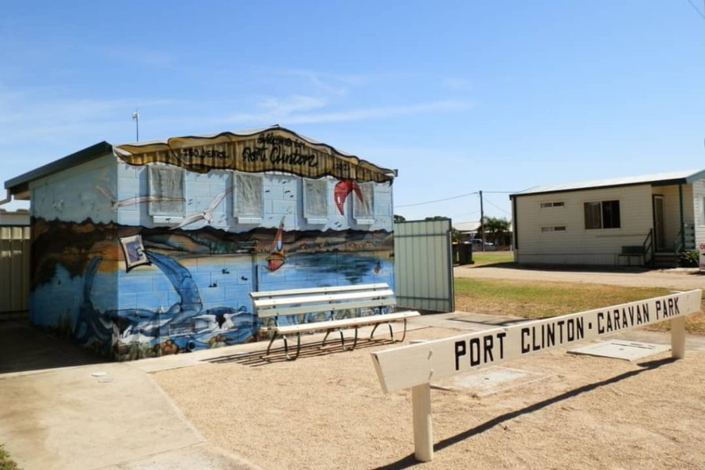 A colorful mural-painted restroom building at Port Clinton Caravan Park in South Australia, featuring seaside-themed artwork with a bench in the foreground and cabins in the background under a sunny sky.