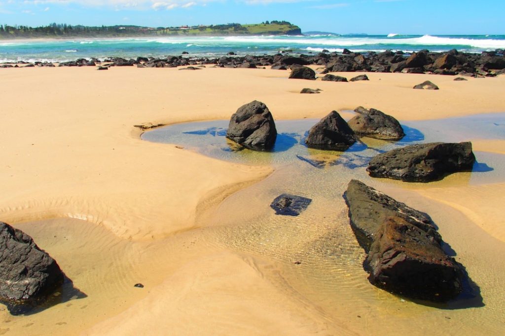 A beach scene with smooth sand, scattered rocks, and shallow pools overlooking Skennars Head, capturing one of the scenic things to do in Ballina NSW for nature lovers.