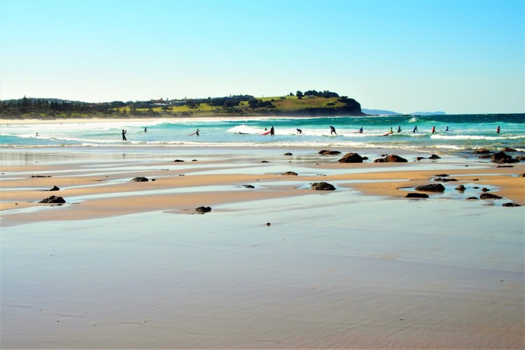Surfers catch waves along the shoreline at Angels Beach with grassy cliffs in the background, highlighting surfing as one of the popular things to do in Ballina NSW.