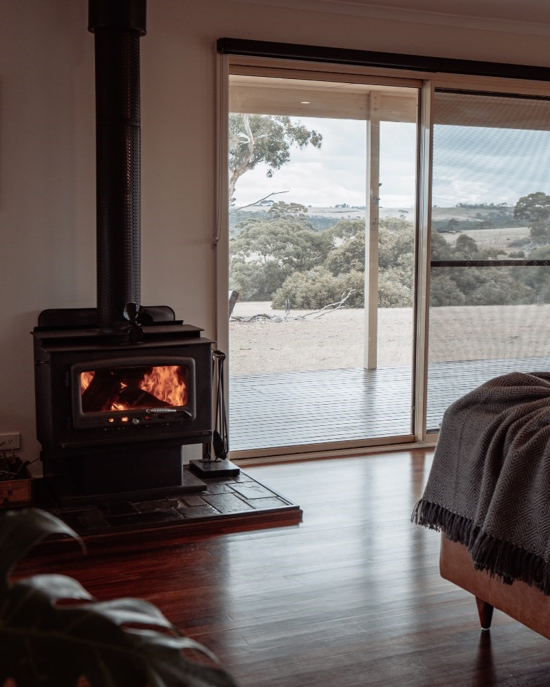A cozy indoor scene at Ecopia Retreat on Kangaroo Island, featuring a wood-burning stove with a warm fire, set against a sliding glass door that opens to a scenic rural landscape with trees and rolling hills. The rustic interior, complete with wooden floors and a blanket-draped bed in the corner, creates an inviting, tranquil atmosphere ideal for relaxation.