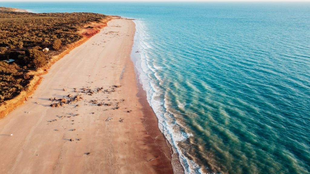 Aerial view of Gantheaume Point, Broome, showing a long stretch of golden sandy beach meeting turquoise ocean waters. The end destination for a road trip from Darwin to Broome