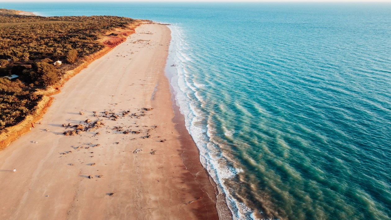 Aerial view of Gantheaume Point, Broome, showing a long stretch of golden sandy beach meeting turquoise ocean waters. The end destination for a road trip from Darwin to Broome