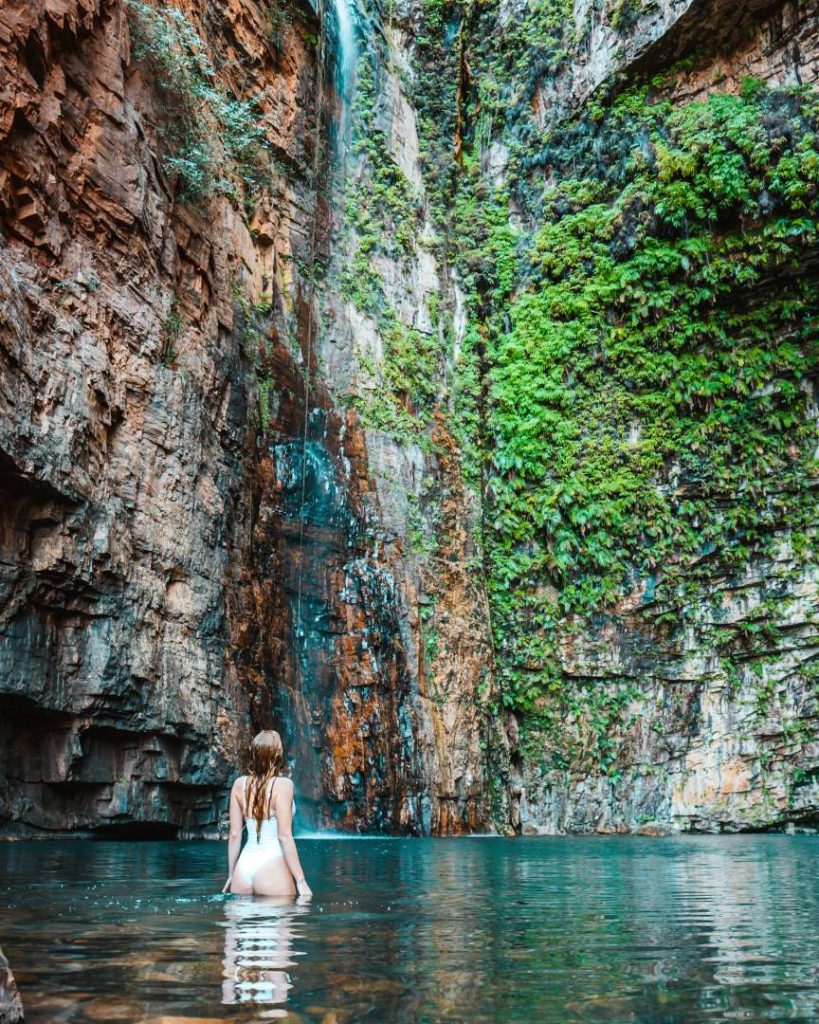 A woman in a swimsuit standing in the crystal-clear pool beneath a tall waterfall surrounded by green ferns and steep rocky cliffs at Emma Gorge, El Questro Station, near Kununurra, Western Australia