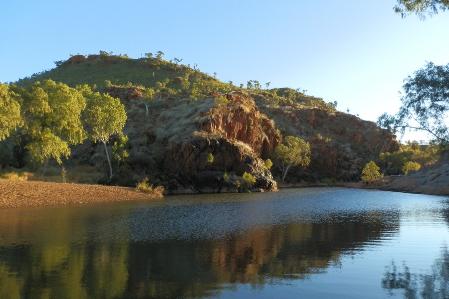 A tranquil pool of water surrounded by red rocky hills and trees under the warm afternoon light at Caroline Pool near Halls Creek.