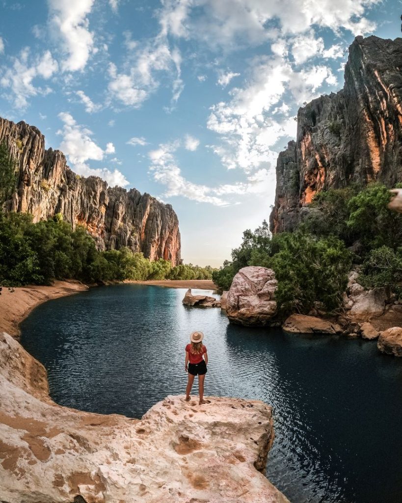 A woman standing on a rock overlooking the turquoise waters and towering cliffs of Windjana Gorge under a partly cloudy sky.